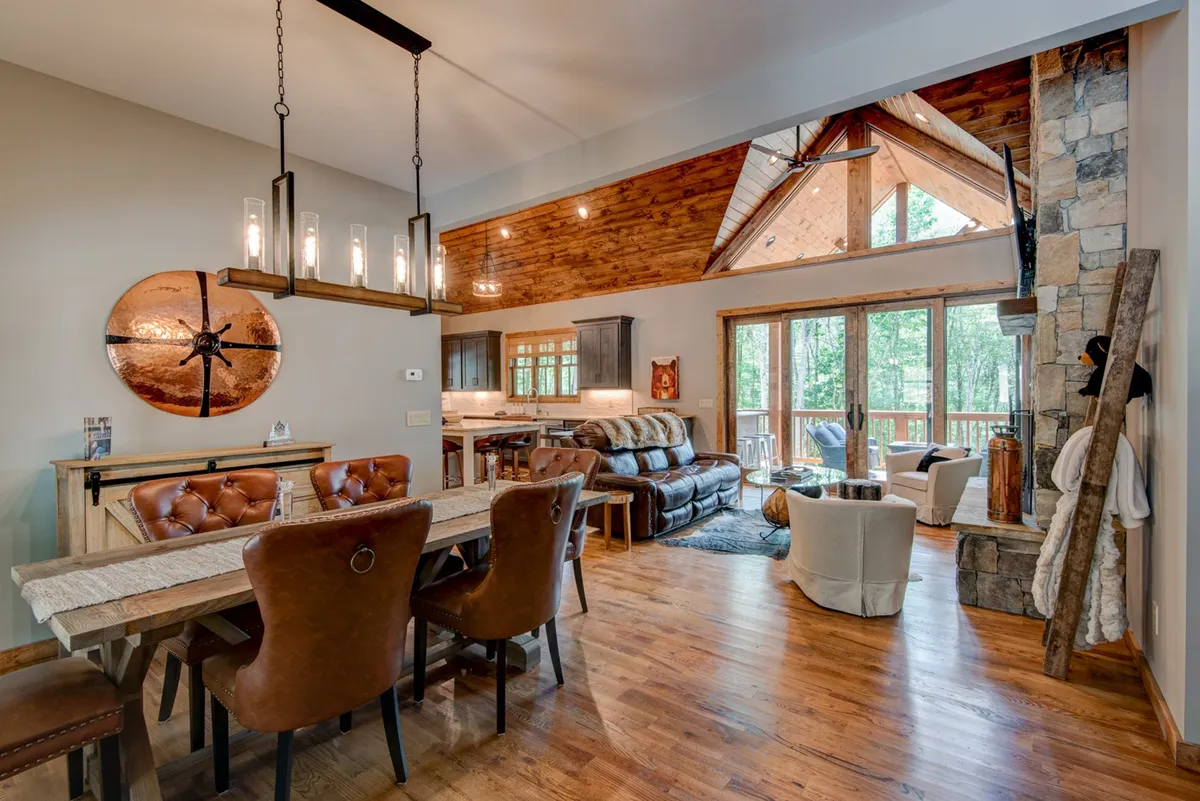 A spacious dining area with a rustic dining table, leather chairs, a chandelier, large windows offering forest views, and a stone fireplace. The layout flows naturally, highlighting the open-concept connection to both gathering and dining spaces.