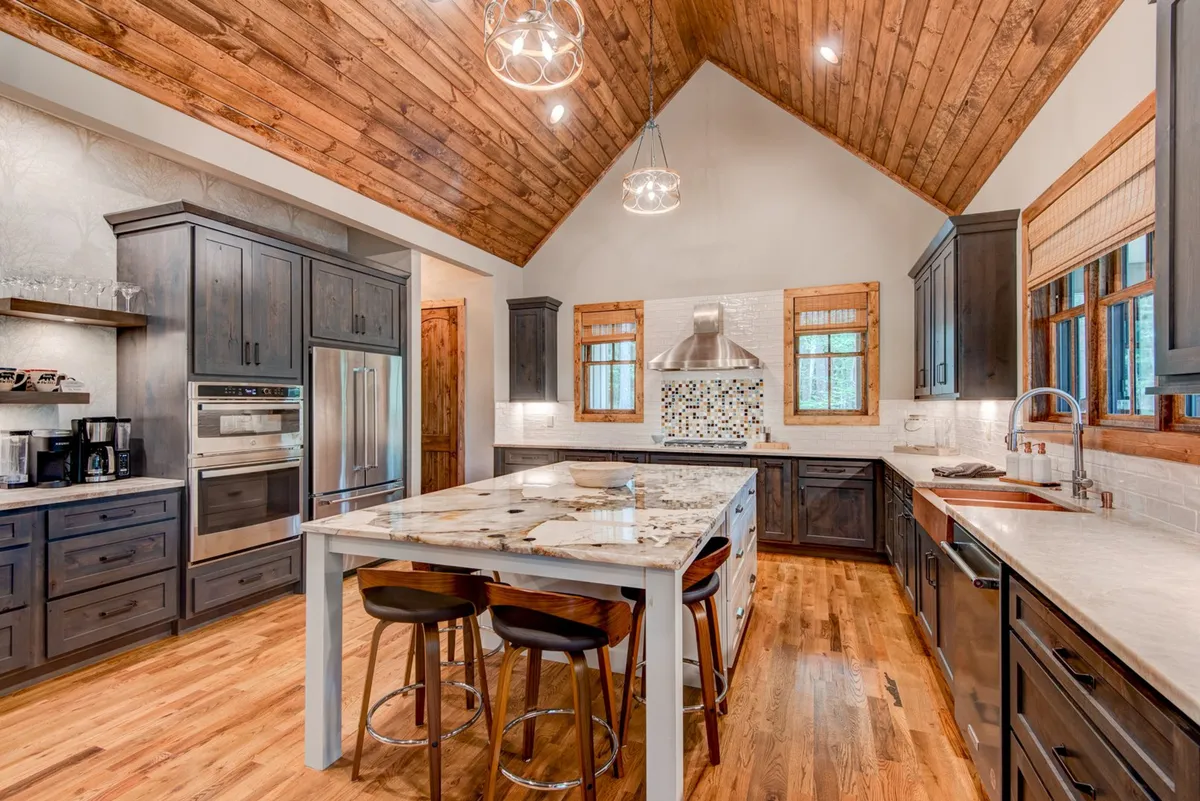 A spacious kitchen featuring a large island topped with luxe stone countertops, modern stainless steel appliances, a decorative backsplash, and a warm wooden ceiling. Natural light brightens the space, highlighting areas perfect for meal prep, socializing, and gathering.