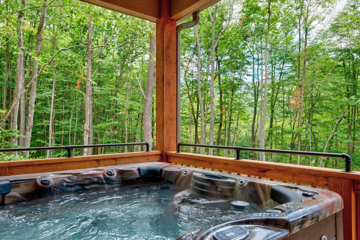 A bubbling hot tub sits on a wooden deck nestled among tall trees, with sunset colors filtering through the forest. The inviting setup suggests relaxation, socializing, and immersion in nature—perfect for unwinding after a day of adventure.