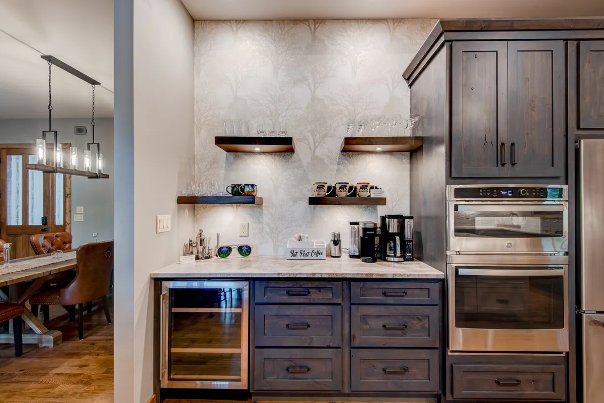 A cozy coffee and cocktail bar setup with dark wooden cabinetry, a state-of-the-art coffee maker, shelves stocked with mugs and glassware, and a warm, inviting ambiance ideal for early mornings or evening drinks.