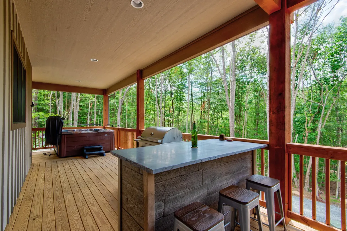 This image showcases a spacious outdoor deck featuring a hot tub, an outdoor grill, a wooden bar counter, and views of surrounding trees. The setting is designed for unwinding, socializing, and enjoying the natural environment, with ample space for group gatherings or quiet conversation.