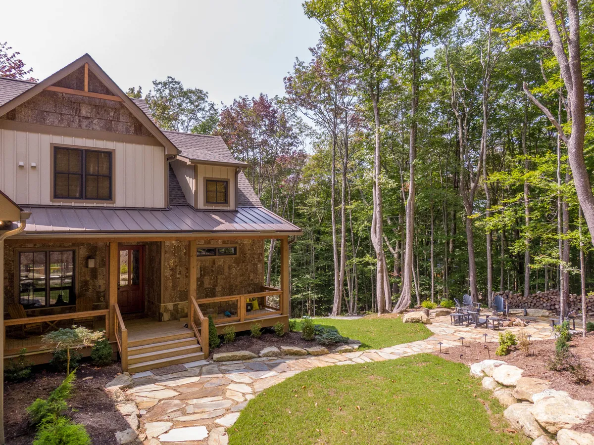 This exterior image highlights a welcoming front porch with comfortable seating, a stone pathway, and mature trees surrounding the area. The inviting porch offers views of a tranquil, natural landscape with a designated fire pit area perfect for evening gatherings.