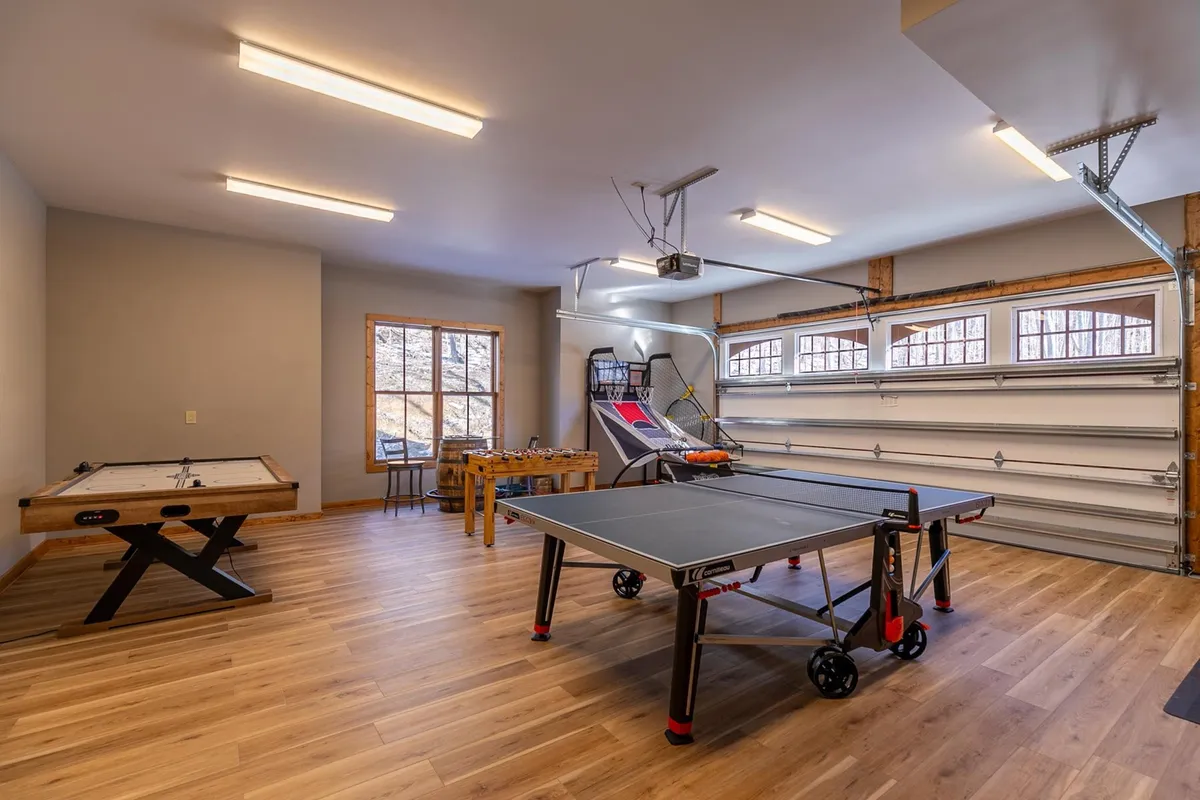 A vibrant and energetic game room featuring a ping pong table, foosball table, and a basketball arcade game, all illuminated by natural light from a large window. The space is designed for friendly competitions and playful family tournaments.
