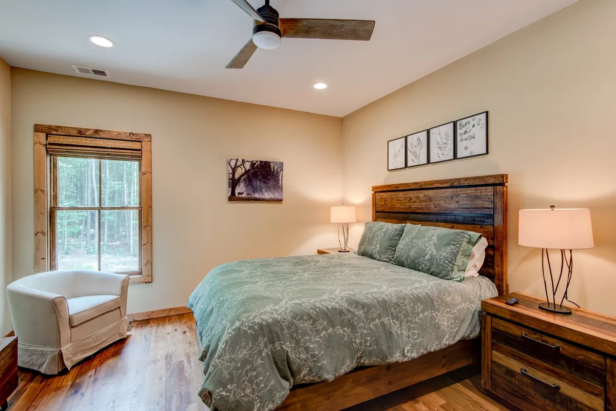A serene bedroom featuring a bed with green bedding, wooden furniture, a large window offering a forest view, and artwork on the wall. Natural light pours in, highlighting the cozy, inviting atmosphere and connection to the outdoors.