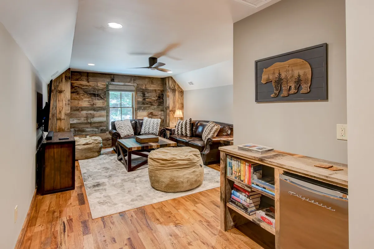 A cozy, inviting upstairs movie and game room with a wood accent wall, leather sofa, coffee table, and bear art piece. The layout encourages movie nights, relaxed gaming, and intimate gatherings.