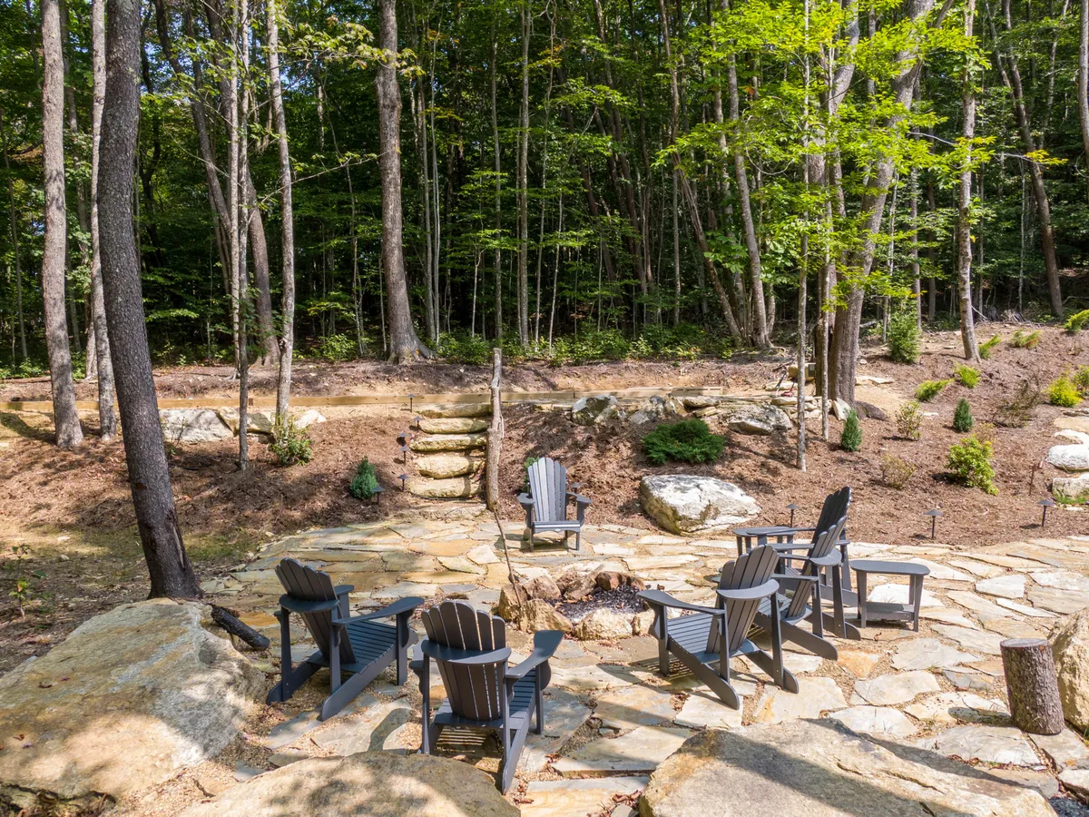 This exterior scene features a cozy fire pit surrounded by Adirondack chairs, set amidst trees and connected by a stone pathway. The arrangement invites groups to gather for storytelling, stargazing, and enjoying the outdoors together during the day or evening.