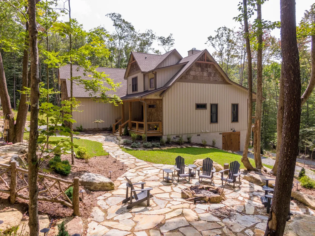 The image features the exterior of a rustic-modern cabin surrounded by lush greenery, with inviting Adirondack chairs arranged around a stone fire pit. A stone pathway leads to a welcoming wooden porch, suggesting ample outdoor space for relaxing, socializing, and enjoying the natural beauty of the mountains.