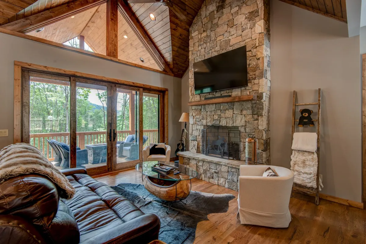 This image showcases a warm, welcoming great room with a prominent stone fireplace, large windows that allow natural light to pour in, a leather sofa, and wooden accents. The arrangement and lighting convey a space ideal for relaxing, socializing, and gathering with friends or family.