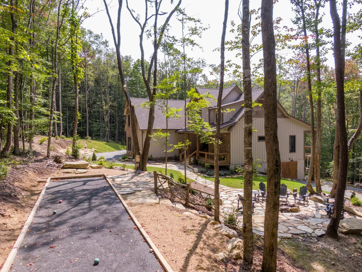 This image showcases the main house with a clear view of the bocce ball court in the foreground, inviting seating areas, and a backdrop of lush, wooded surroundings. The scene is playful and serene, with space for outdoor games, gatherings, and exploration.