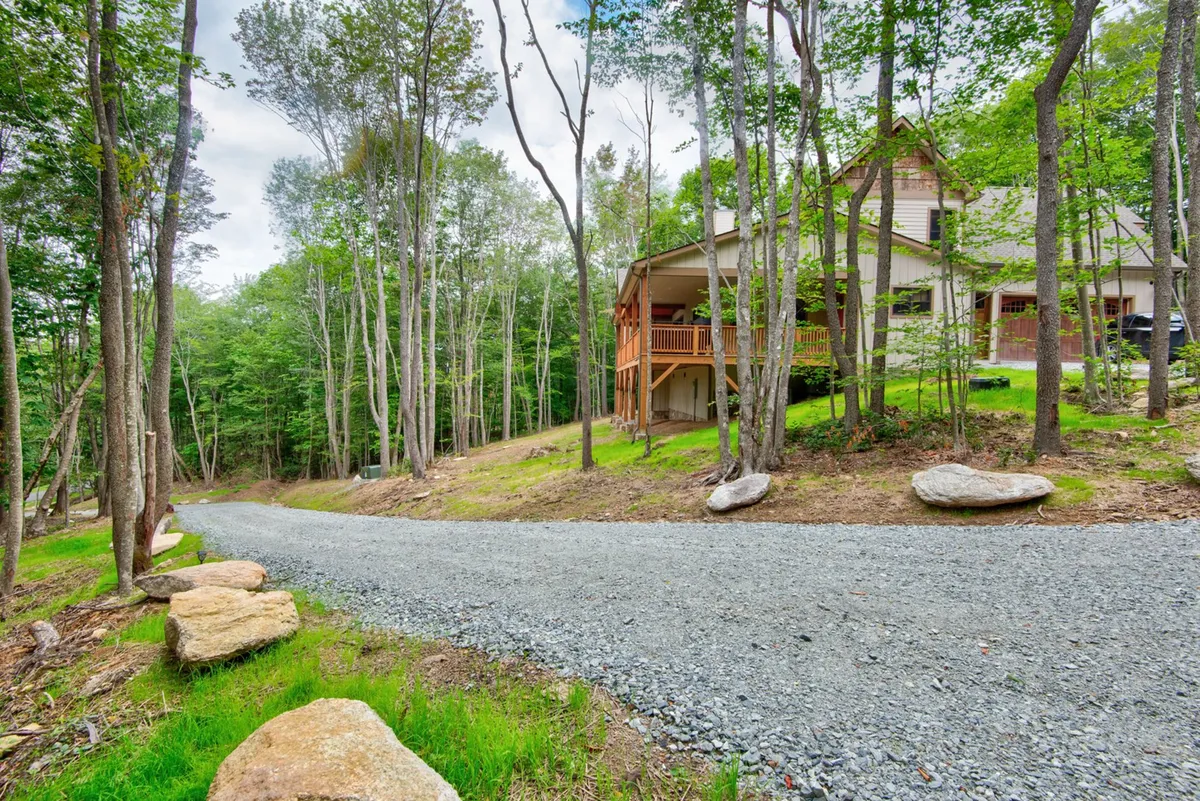This exterior shot features the house nestled among tall trees, with a gravel driveway leading up to the property and a welcoming porch. The surrounding nature and open space are prominent, giving a sense of the acreage and freedom for both people and pets to explore.