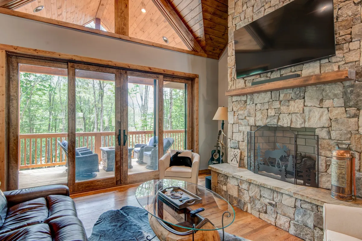A warm, inviting interior with a stone fireplace, comfortable seating area, large glass doors that frame views of the outdoors, and rustic wooden beams. The space is ideal for group meetings, quiet productivity, or unwinding by the fire with nature just outside.