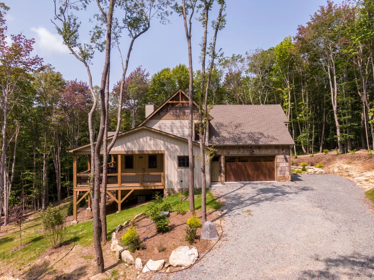 A welcoming front porch with a wooden garage door, surrounded by landscaped gardens and mature trees. The setting hints at both relaxation and the start of adventure, with nearby woods and trails extending beyond the property.