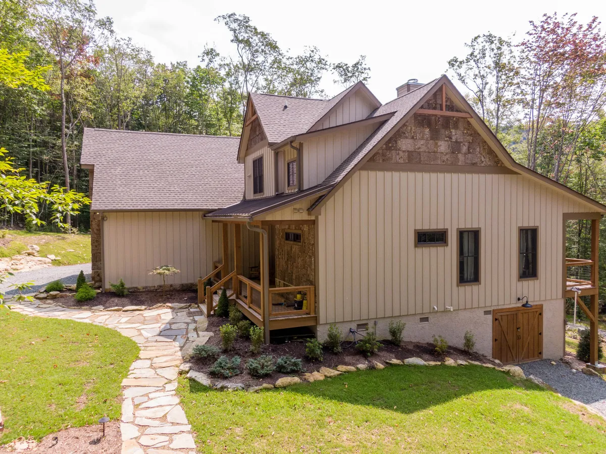 An exterior shot featuring a broad wooden porch, stone pathway, landscaped greenery, and dormer windows under a mountain sky. The porch is spacious enough for group activities or quiet coffee breaks, while the surrounding trees and landscaping enhance the serene retreat vibe.