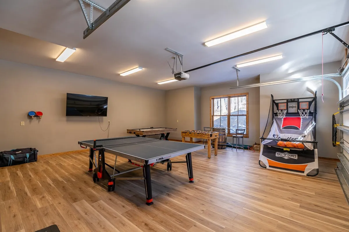 A vibrant, energetic garage game room featuring a ping pong table, foosball table, basketball arcade game, and wall-mounted TV. The setup is designed for family tournaments and playful competitions, offering a fun, dynamic vibe.